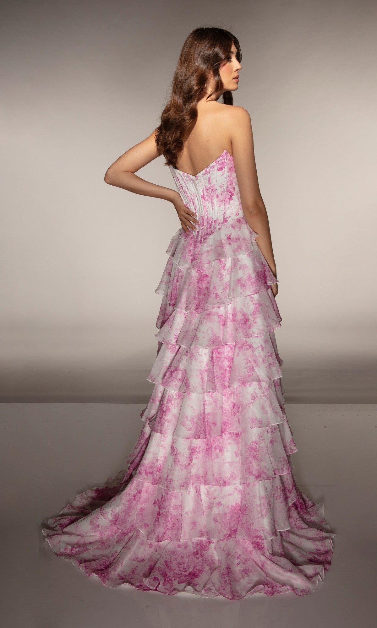 A woman with long brown hair, seen from the back, wears the Alyce 62192 long prom dress featuring pink floral patterns and a strapless chiffon ruffle skirt.